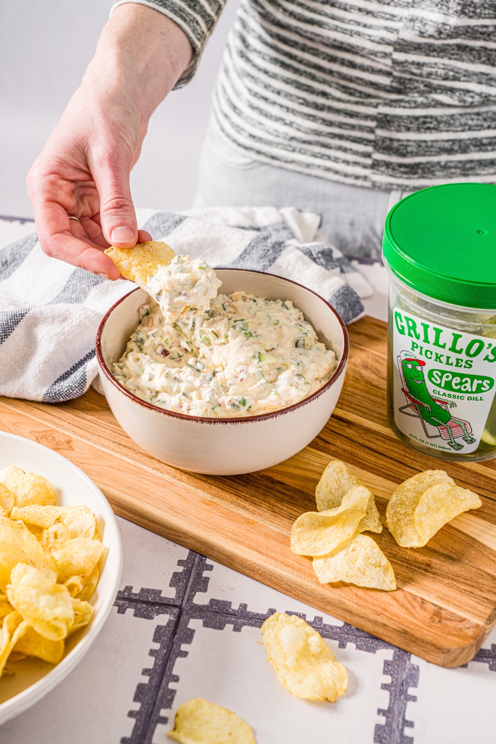 A hand holding a potato chip that has Grillo's pickle dip on it being held over a bowl of the creamy pickle dip. To the right of that bowl is a tub of Grillo's pickles, and both are on top of a wooden cutting board.
