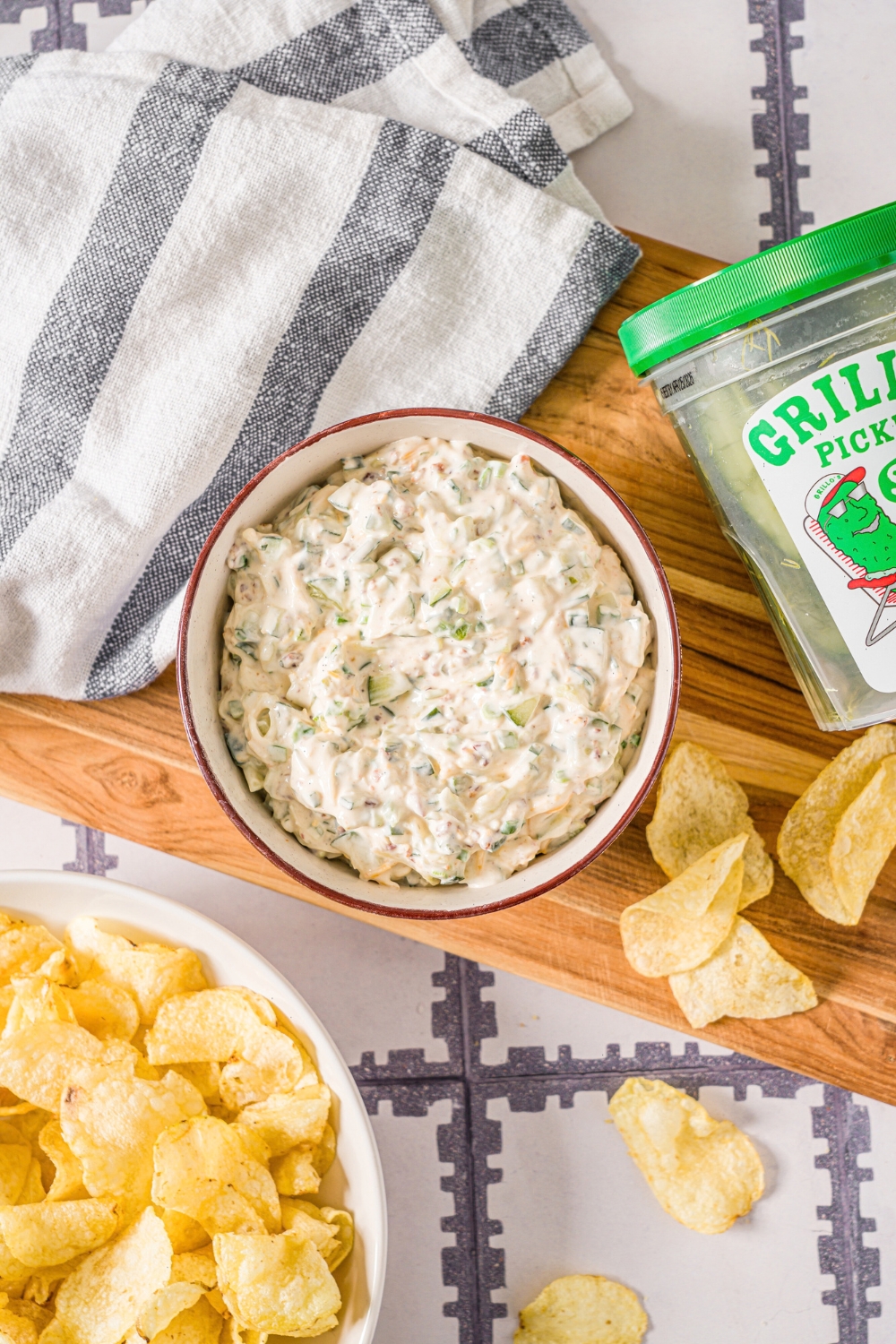 A creamy grilled pickle dip in a white bowl on top of a wooden cutting board with a tub of Grillo's pickles next to the dip and some potato chips on the cutting board as well.