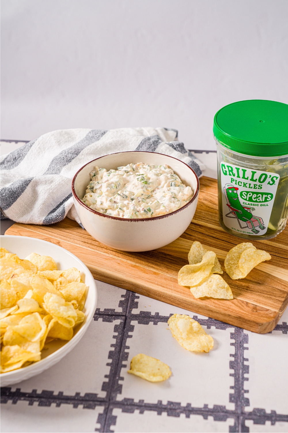 A tub of Grillos pickles with a white bowl to the left of it that is filled with a creamy pickle dip. They are on top of a wooden cutting board and in front of the cutting board is part of a bowl with potato chips in it.