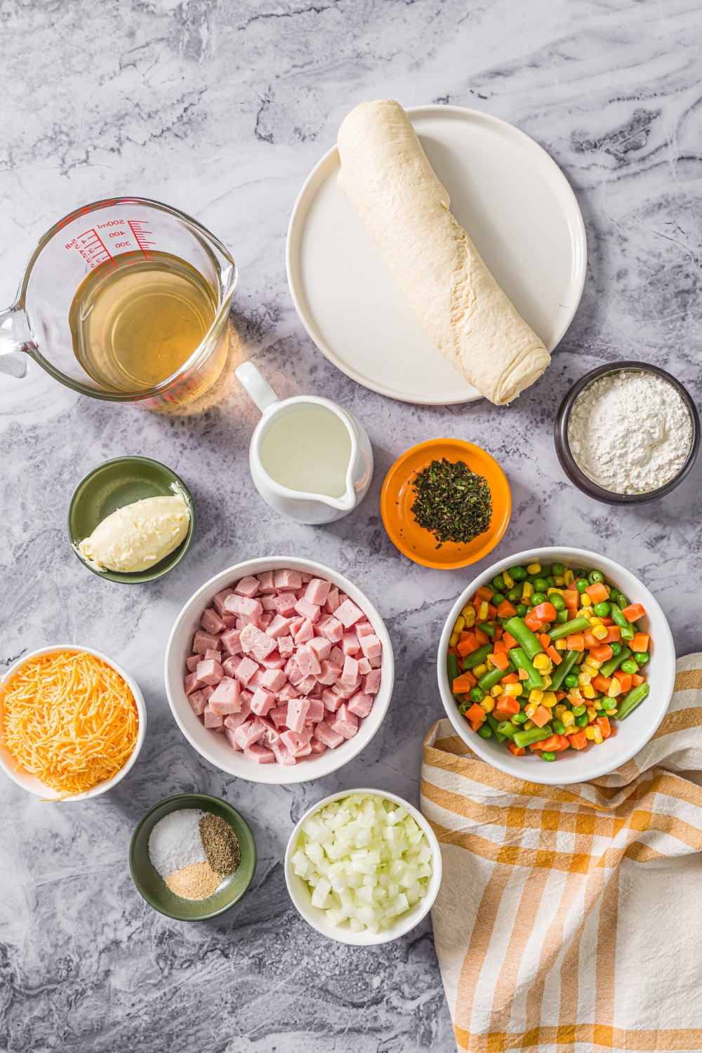 A marble counter with several bowls in various sizes containing ingredients to make ham pot pie including cubed ham, mixed vegetables, flour, onion, crescent dough, chicken stock, heavy cream, shredded cheddar cheese, and seasonings.