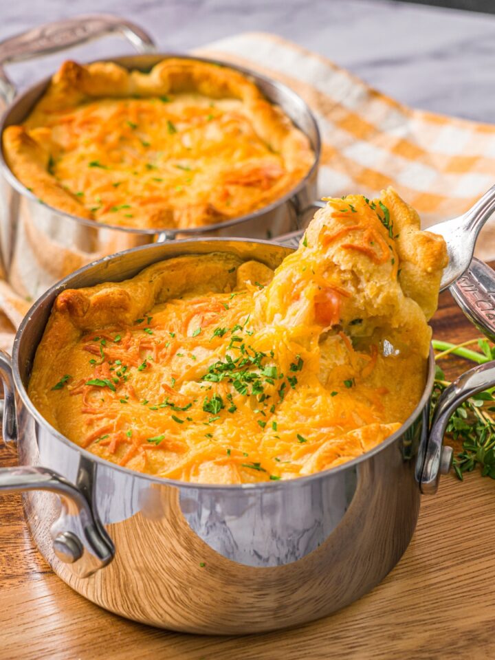 A small pie pot with baked ham pot pie garnished with fresh parsley. There is a fork taking a bite of the pie. The pot is on a wooden board. The board is on a marble counter with a yellow checkered napkin with an additional pot pie.