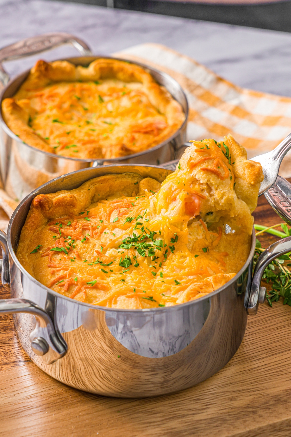 A small pie pot with baked ham pot pie garnished with fresh parsley. There is a fork taking a bite of the pie. The pot is on a wooden board. The board is on a marble counter with a yellow checkered napkin with an additional pot pie.