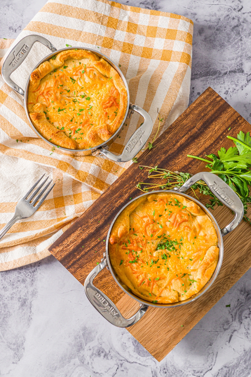 A marble counter with a pie pot with baked ham pot pie garnished with fresh parsley. One pot is on a wooden board and the other is on a yellow checkered napkin.