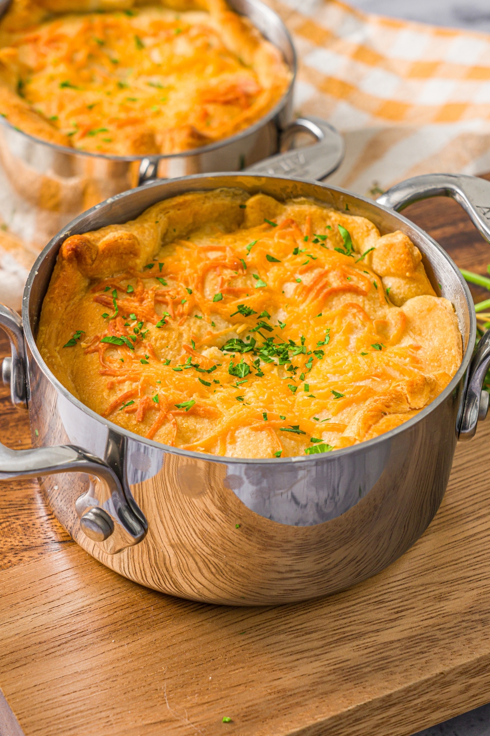 A pie pot with a baked ham pot pie garnished with fresh parsley. The pot is on a wooden board on a marble counter with an additional pot pie with a yellow checkered napkin.