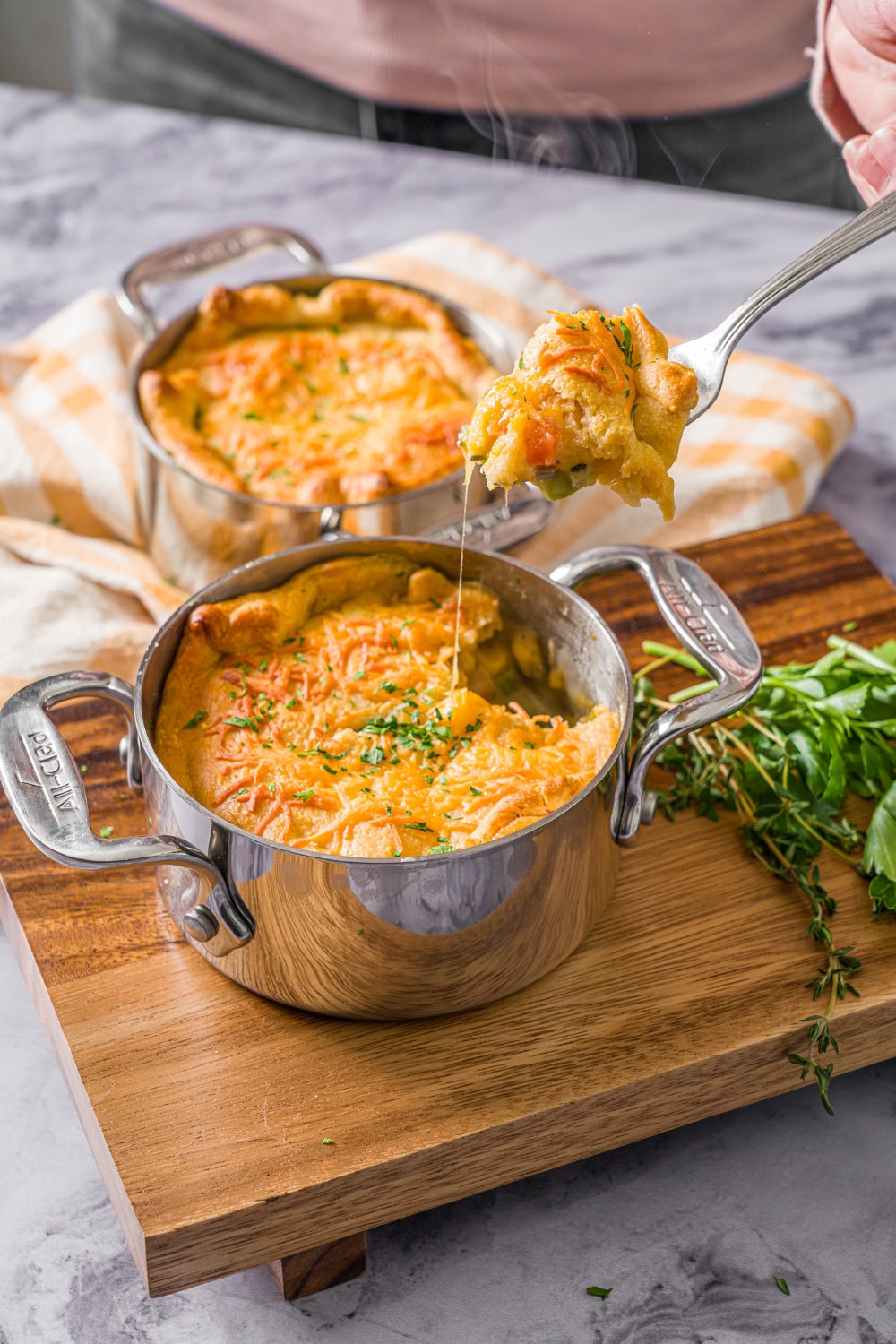 A pie pot with a baked ham pot pie garnished with fresh parsley. A fork is taking a bite from the pie. The pot is on a wooden board on a marble counter with an additional pot pie with a yellow checkered napkin.