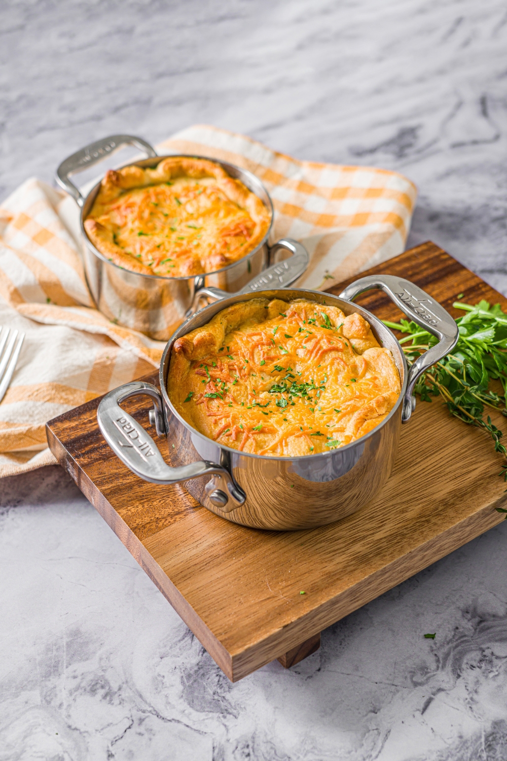 A pie pot with a baked ham pot pie garnished with fresh parsley. A fork is taking a bite from the pie. The pot is on a wooden board on a marble counter with an additional pot pie on a yellow checkered napkin.