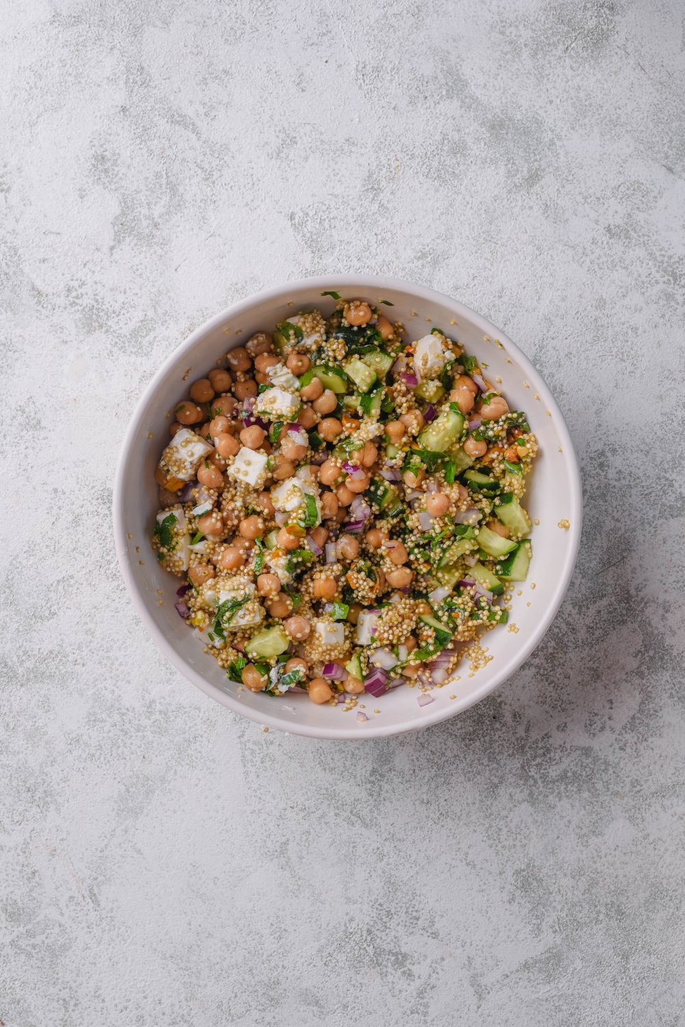 A white bowl with ingredients mixed to make Jennifer Aniston salad including quinoa, cucumber, red onion, feta cheese, chick peas, roasted pistachios, and mint tossed in lemon dressing. The bowl is on a marble counter.