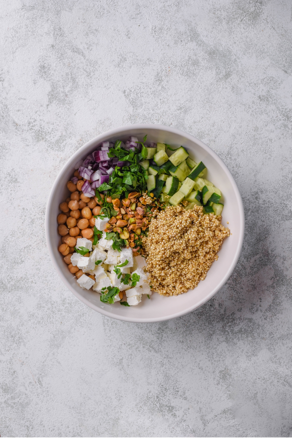A white bowl with ingredients to make Jennifer Aniston salad including quinoa, cucumber, red onion, feta cheese, chick peas, roasted pistachios, and mint. The bowl is on a marble counter.