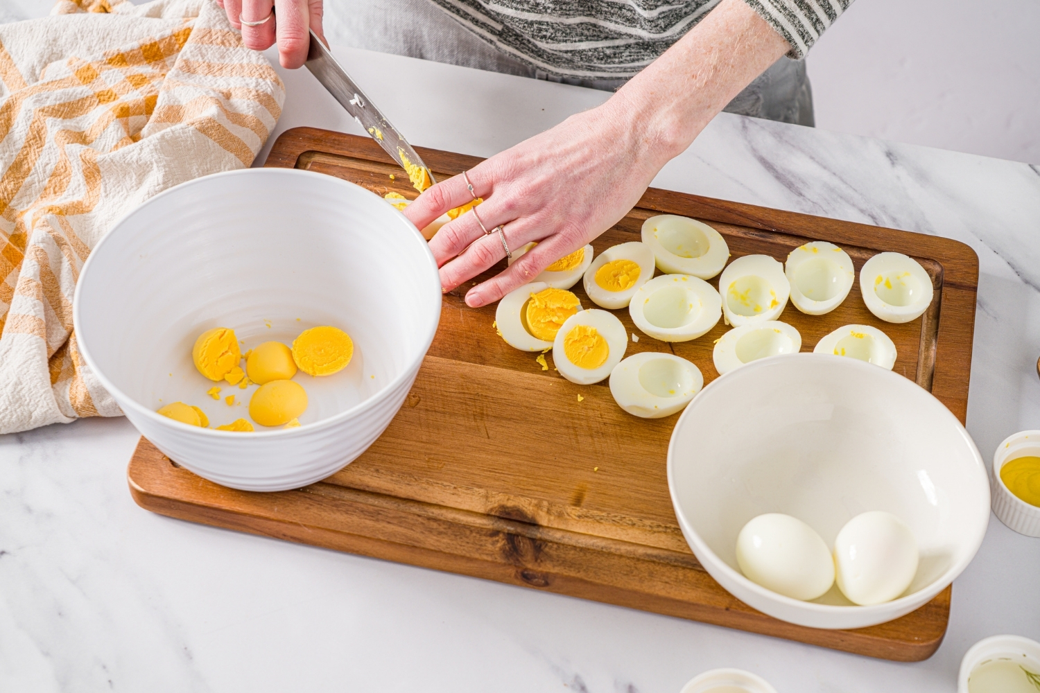 A wooden board with several hard boiled eggs being sliced in half. The yolks are being added to a large white bowl. The board is on a marble counter.