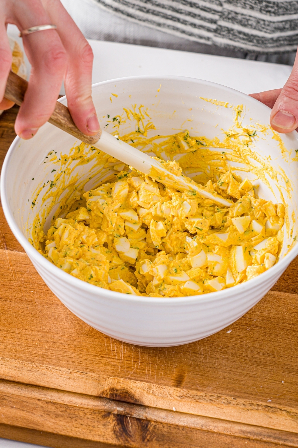 A large white bowl with a spatula stirring an egg salad mixture. The bowl is on a wooden board.