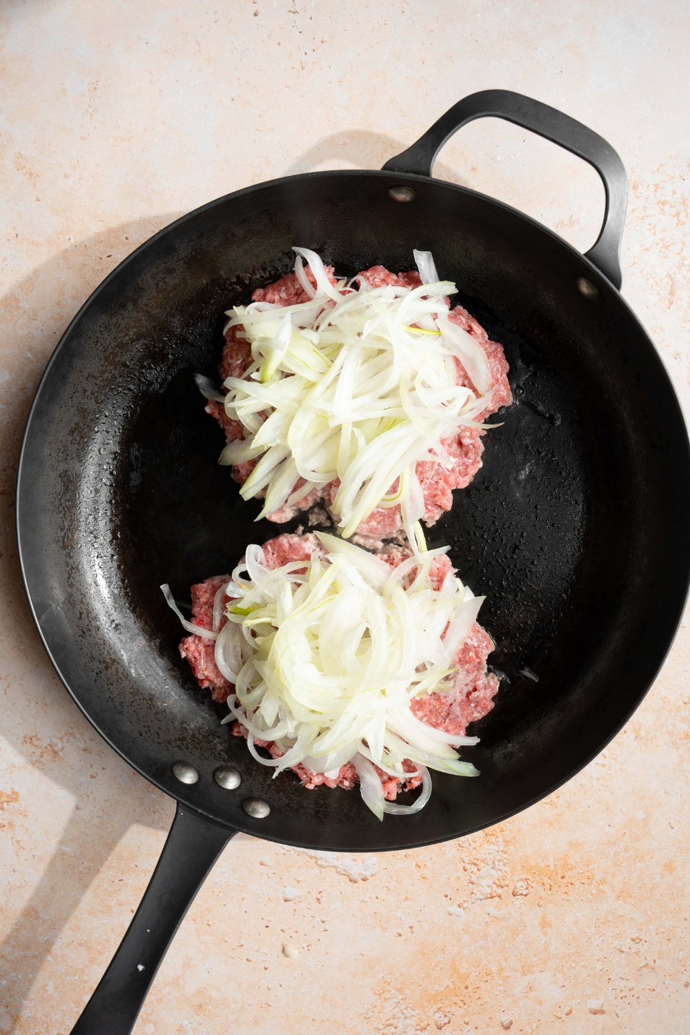 A skillet with two smash burger patties cooking with onion. The skillet is on a tan counter.
