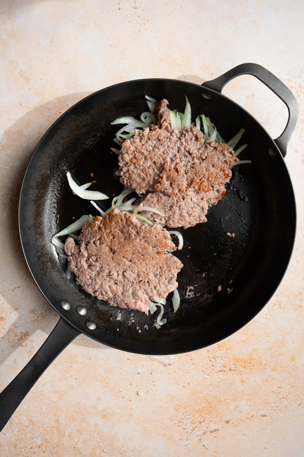 A skillet with two cooked smash burgers with onions. The skillet is on a tan counter.
