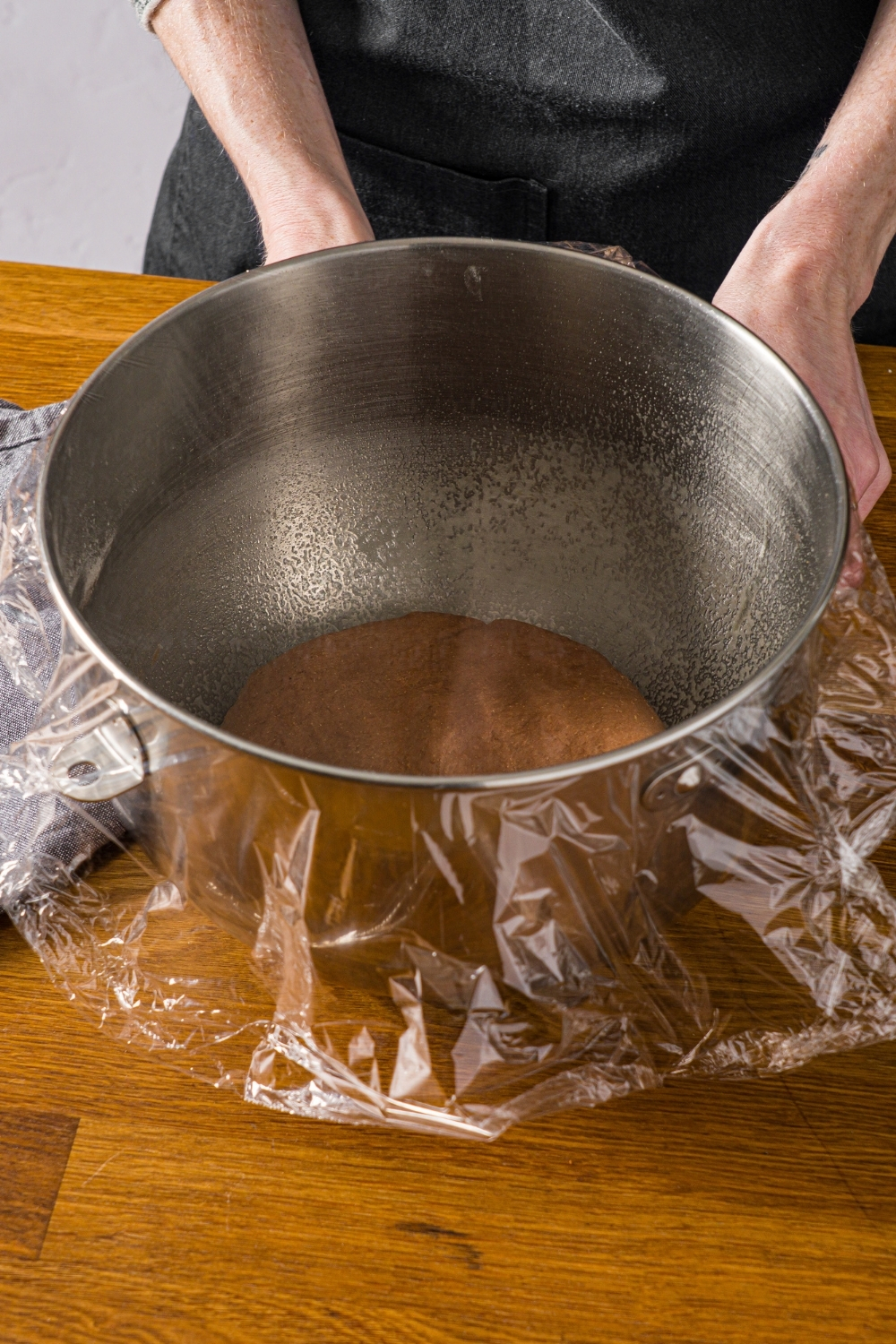 A large mixing bowl with Outback sweet molasses brown bread dough covered with plastic wrap. The bowl is on a wooden counter.