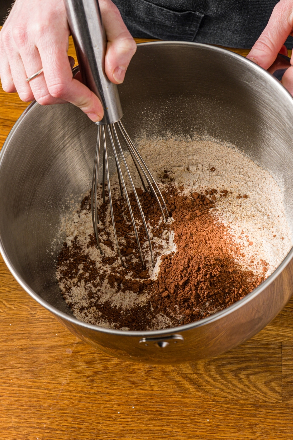 A large mixing bowl with dry ingredients to make Outback sweet molasses brown bread including flour, cocoa powder, yeast, and more. A whisk is mixing the ingredients.