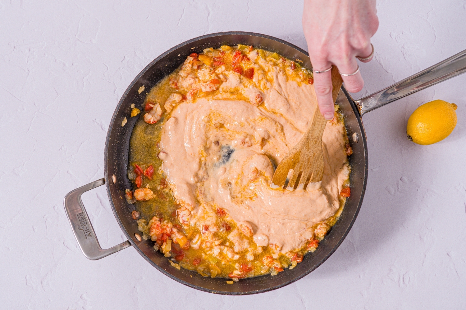 A skillet with sautéed onions, tomatoes, and shrimp with a wooden spoon stirring in Trader Joe's lobster bisque. The skillet is on a marble counter.