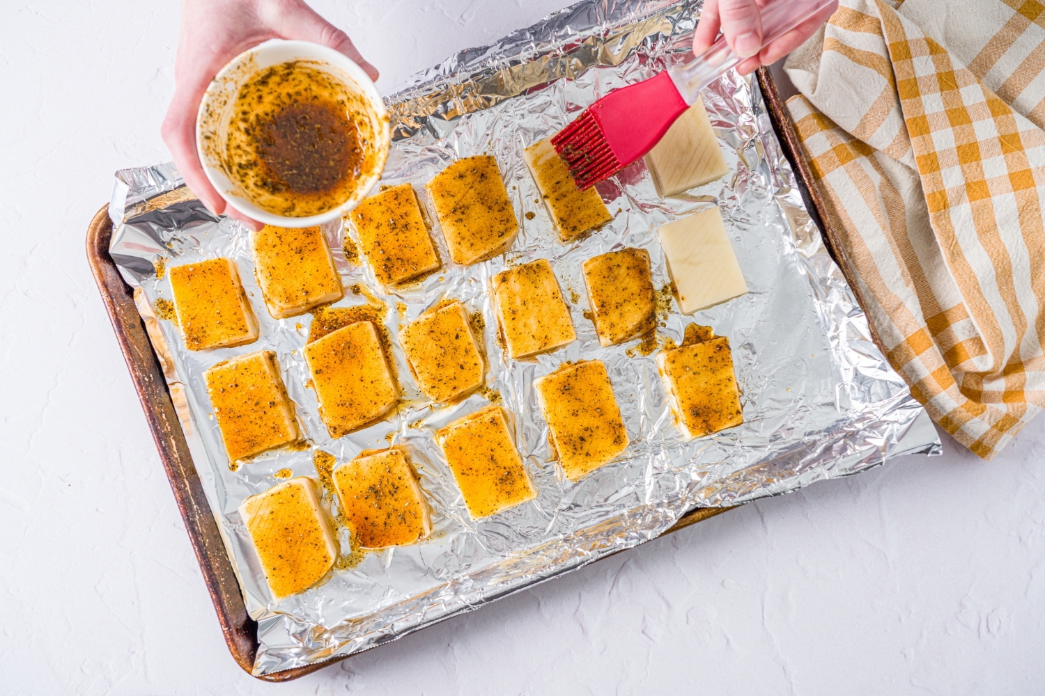 A baking sheet lined with foil with unbaked accordion potatoes. The potatoes are being brushed with an oil and seasoning mixture. The sheet is on a white counter with a yellow checkered napkin.