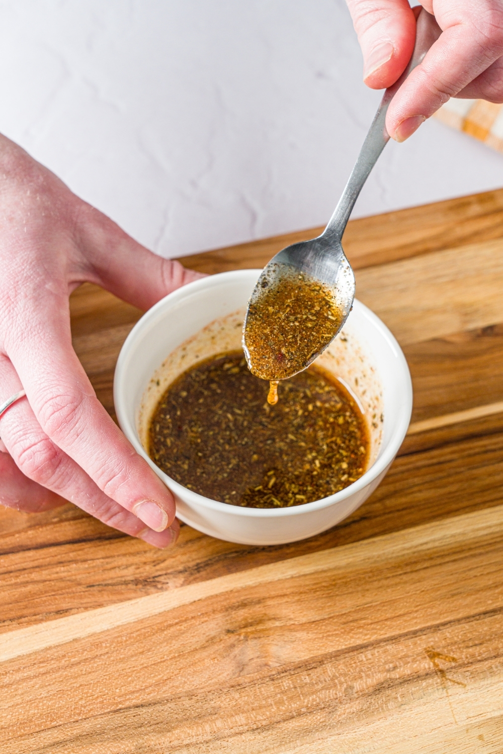 A small white bowl with a spoon mixing an oil and seasonings mixture. The bowl is on a wooden board.