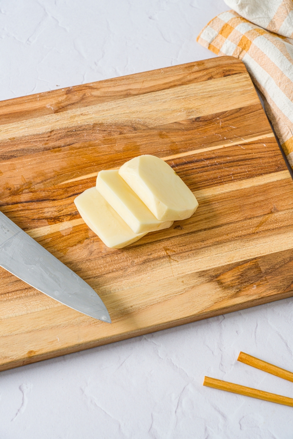 A wooden board with three slices of peeled and trimmed potatoes. The board is on a white counter with a yellow checkered napkin.