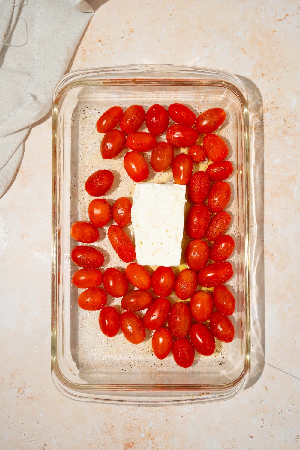 A glass baking dish with fresh cherry tomatoes and a block of feta cheese. The dish is on a tan counter with a white cloth napkin.