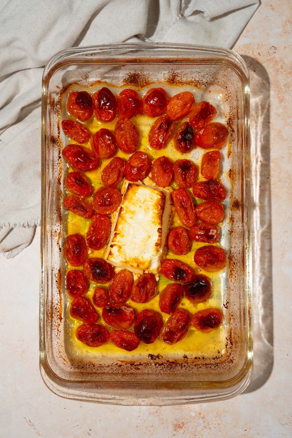 A glass baking dish with baked cherry tomatoes and a block of feta cheese. The dish is on a tan counter with a white cloth napkin.