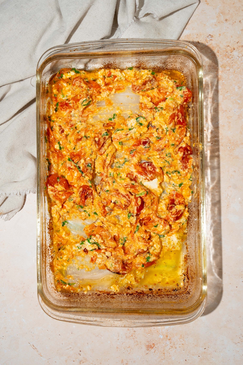 A glass baking dish with baked cherry tomatoes, feta cheese, and fresh parsley mixed together. The dish is on a tan counter with a white cloth napkin.