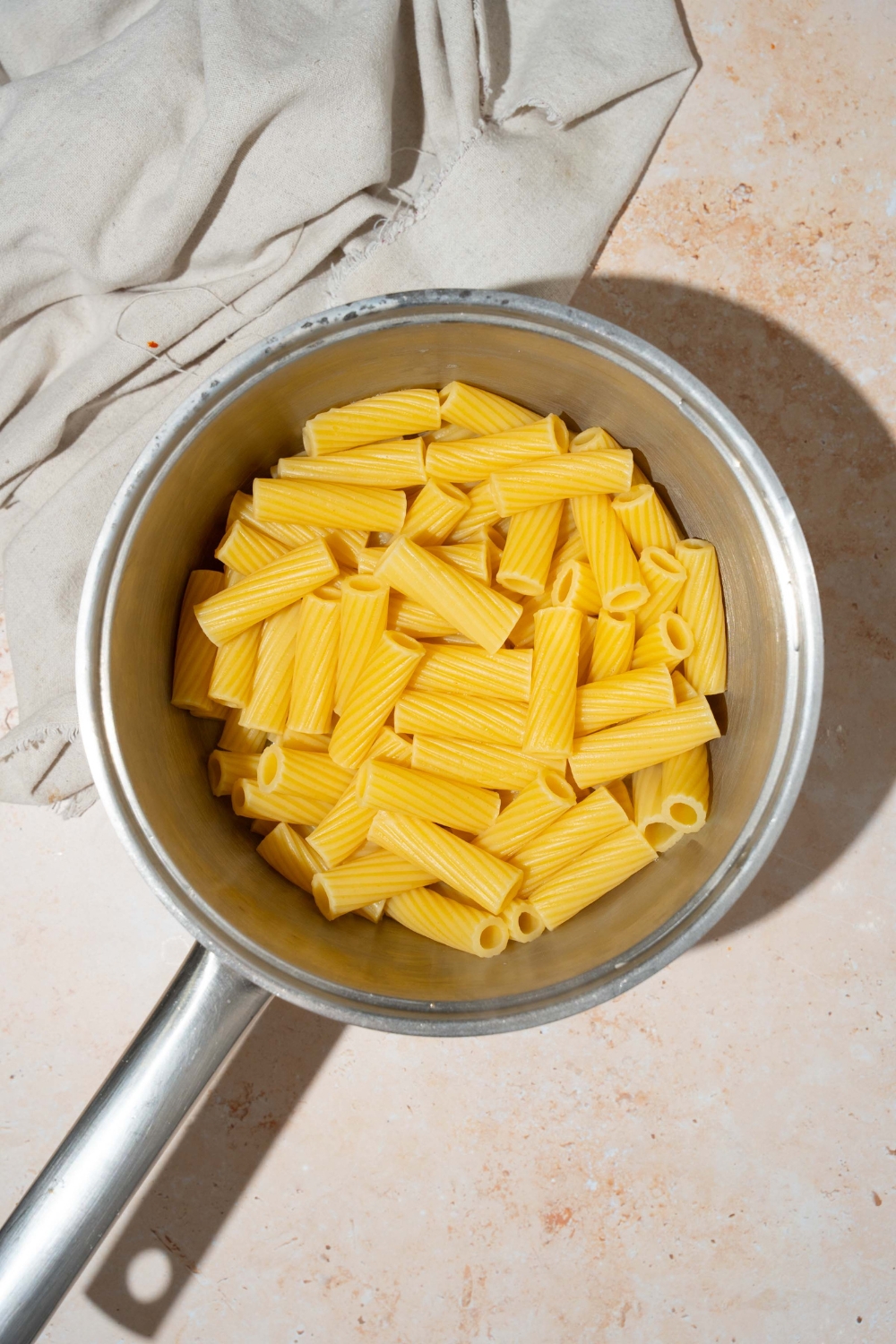 A pot with cooked rigatoni pasta. The pot is on a tan counter with a white cloth napkin.