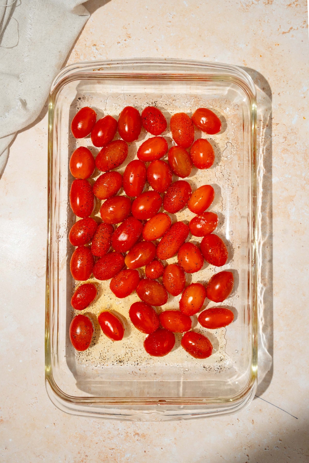 A glass baking dish with cherry tomatoes drizzled with olive oil. The dish is on a tan counter with a white cloth napkin.