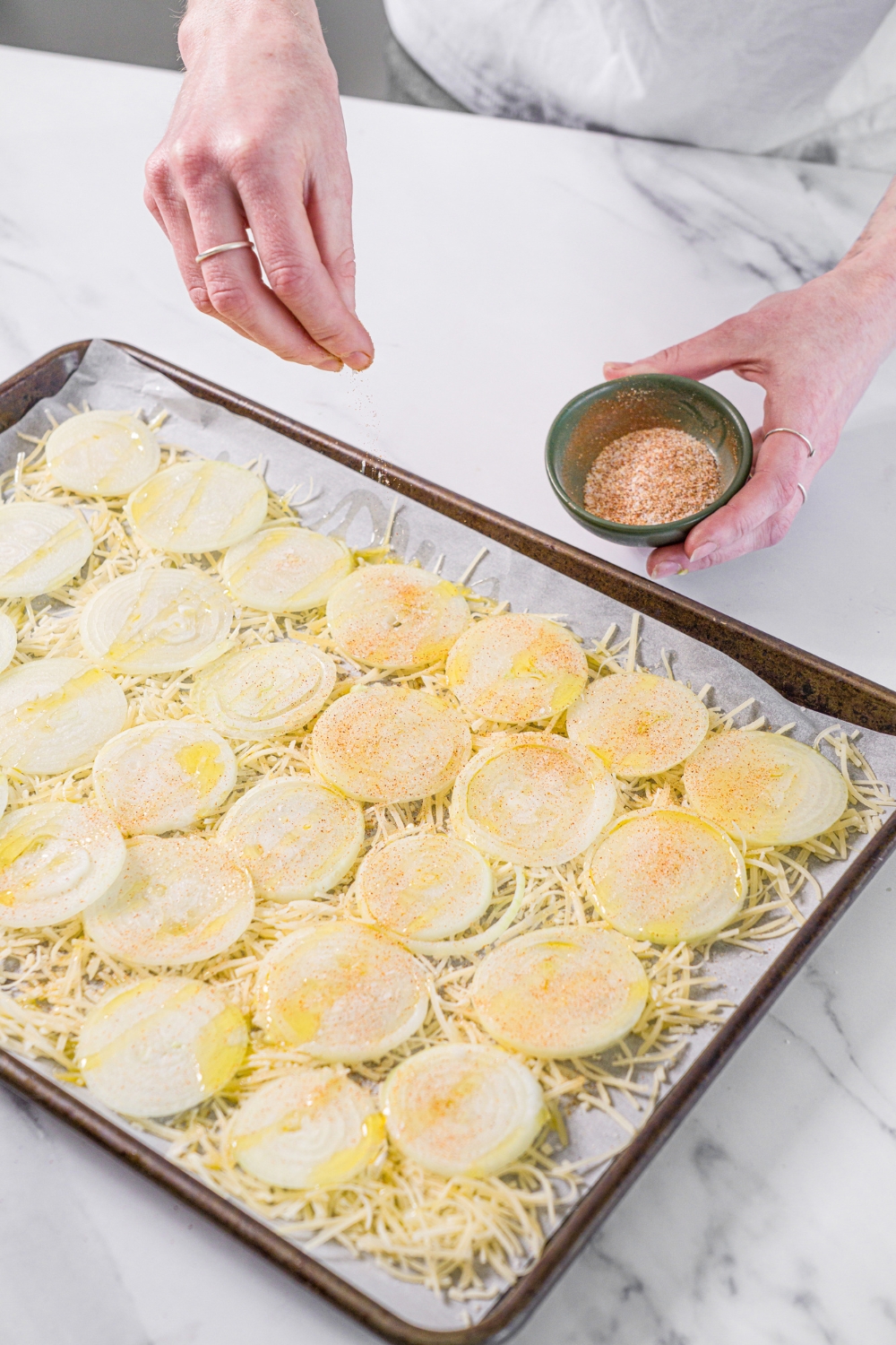 A baking sheet lined with parchment paper with shredded parmesan cheese topped with sliced onions. The onions are drizzled with oil and seasonings are being added to the onions. The sheet is on a marble counter.