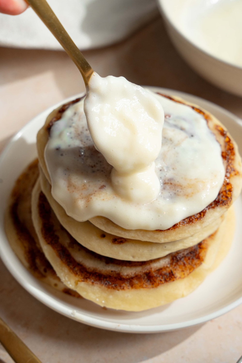 A white plate with a stack of cinnamon roll pancakes with a spoon pouring cream cheese icing over the stack. The plate is on a tan counter.