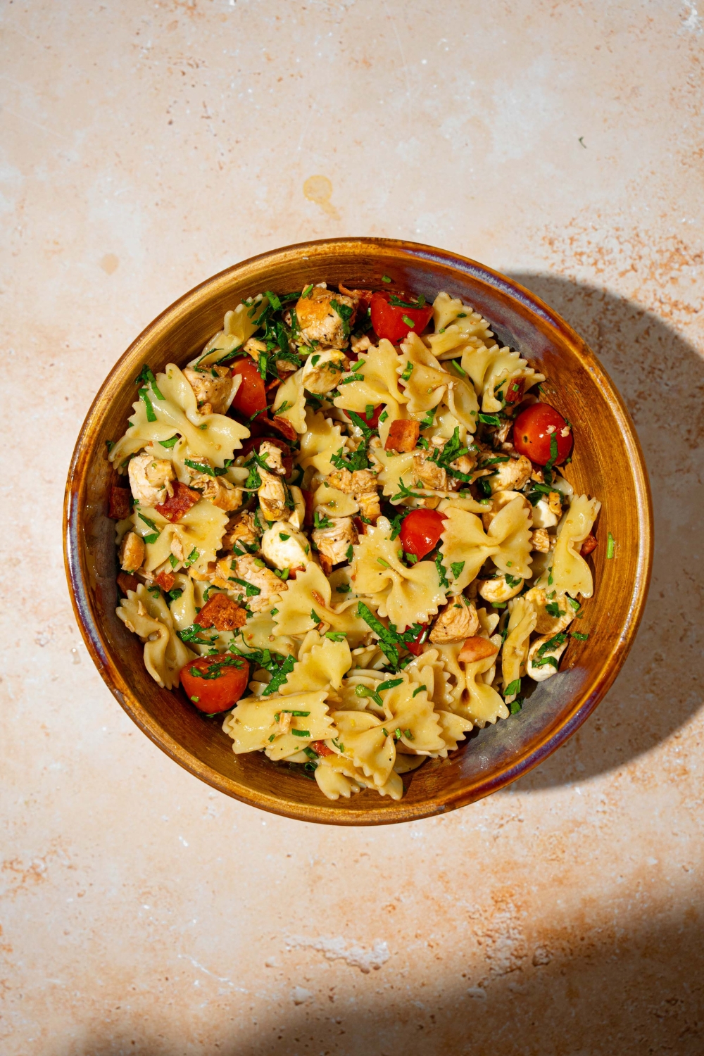 A large bowl with bowtie pasta, tomatoes, mozzarella pearls, fresh basil, bacon, and chicken tossed in a balsamic vinaigrette. The bowl is on a tan counter.