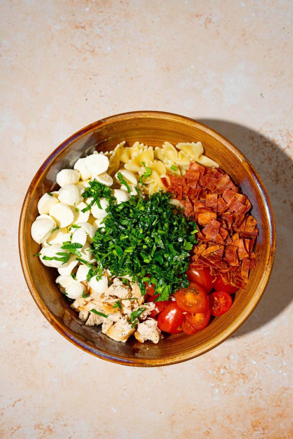 A large bowl with ingredients to make bruschetta chicken pasta including bowtie pasta, mozzarella pearls, tomatoes, fresh basil, and bacon. The bowl is on a tan counter.
