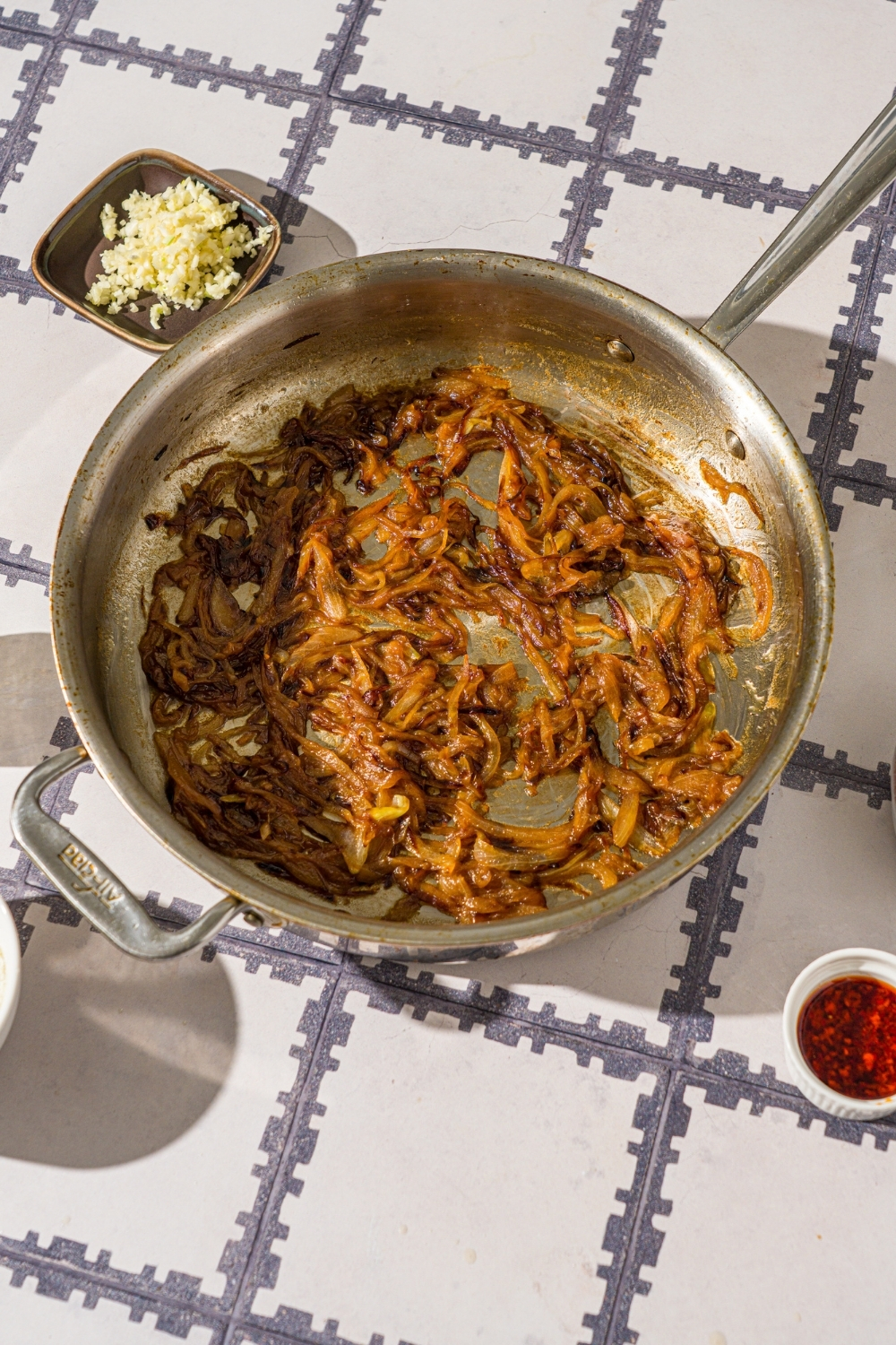 A skillet with caramelized onions. The skillet is on a tiled counter with a bowl of garlic and chili crisp oil.