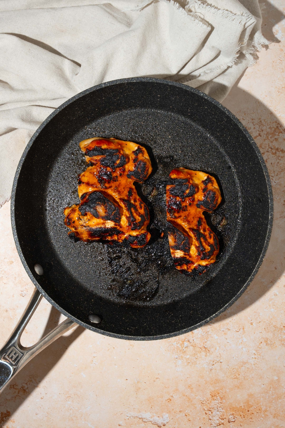A skillet with grilled marinated chicken thighs. The skillet is on a tan counter with a white cloth napkin.