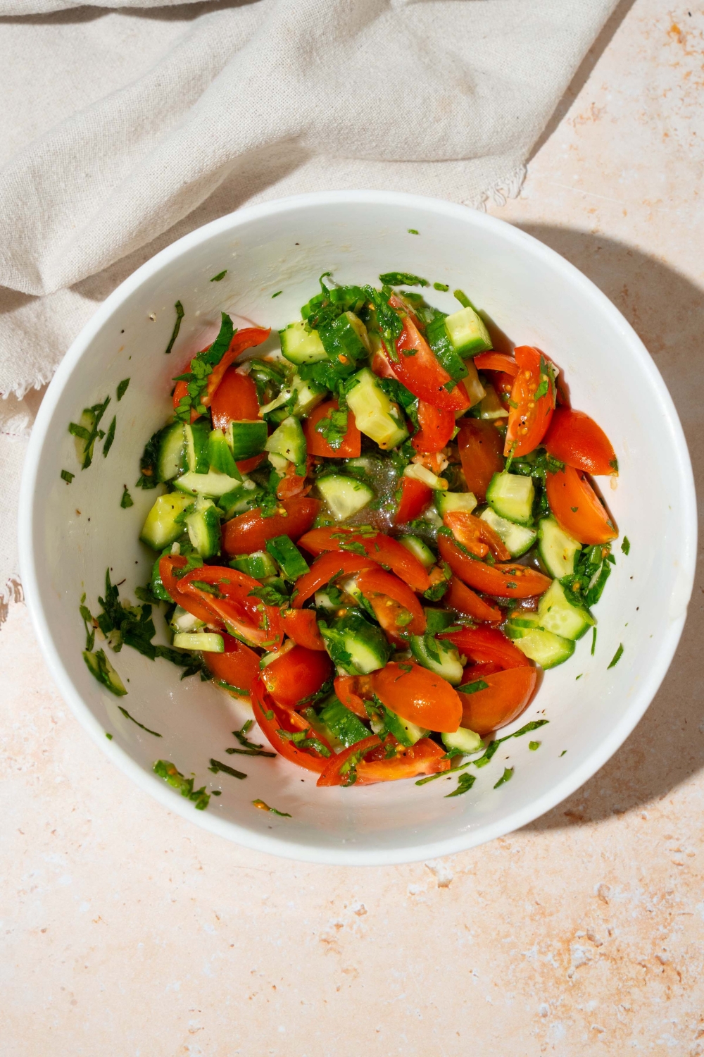 A white bowl with a cucumber and tomato salad tossed in oil. The bowl is on a tan counter with a white cloth napkin.