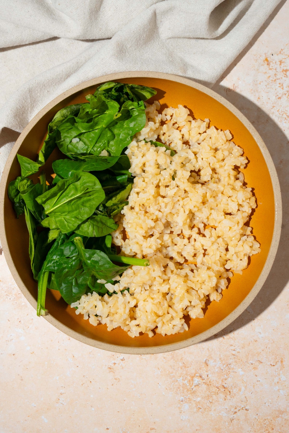 A bowl with cooked rice and mixed greens. The bowl is on a tan counter with a white cloth napkin.