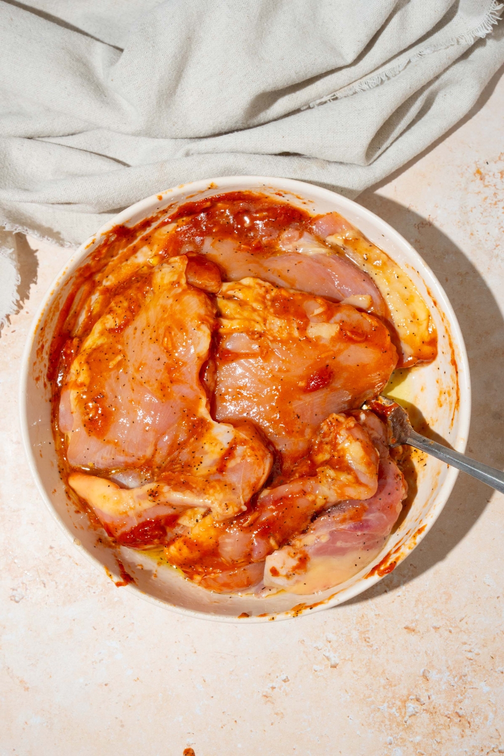 A large white bowl with chicken thighs marinating in a honey harissa marinade. The bowl is on a tan counter with a white cloth napkin.