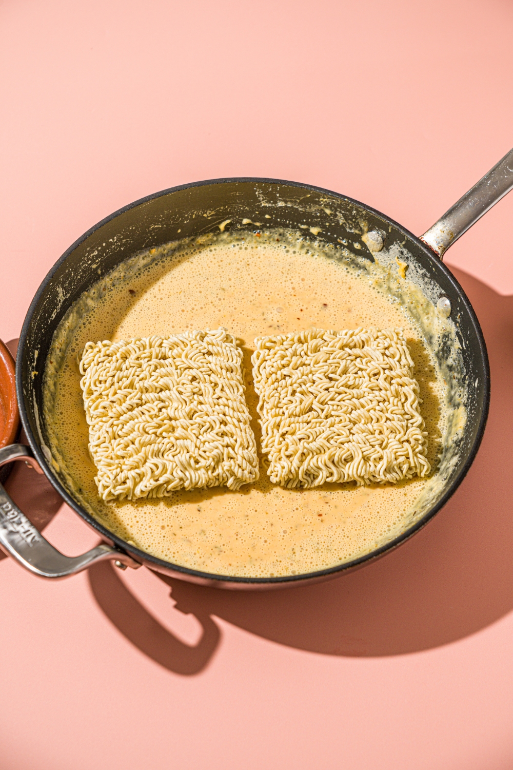 A skillet with maggi noodles sitting in a seasoned cheese sauce. The skillet is on a pink counter.