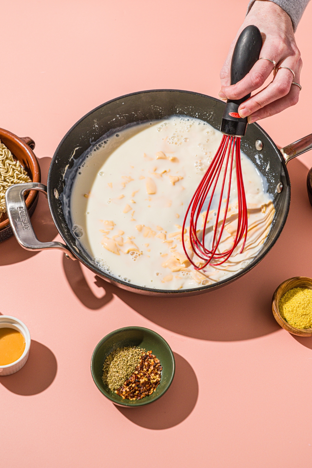 A skillet with a spatula mixing milk and American cheese. The skillet is on a pink counter with small bowls of ingredients.