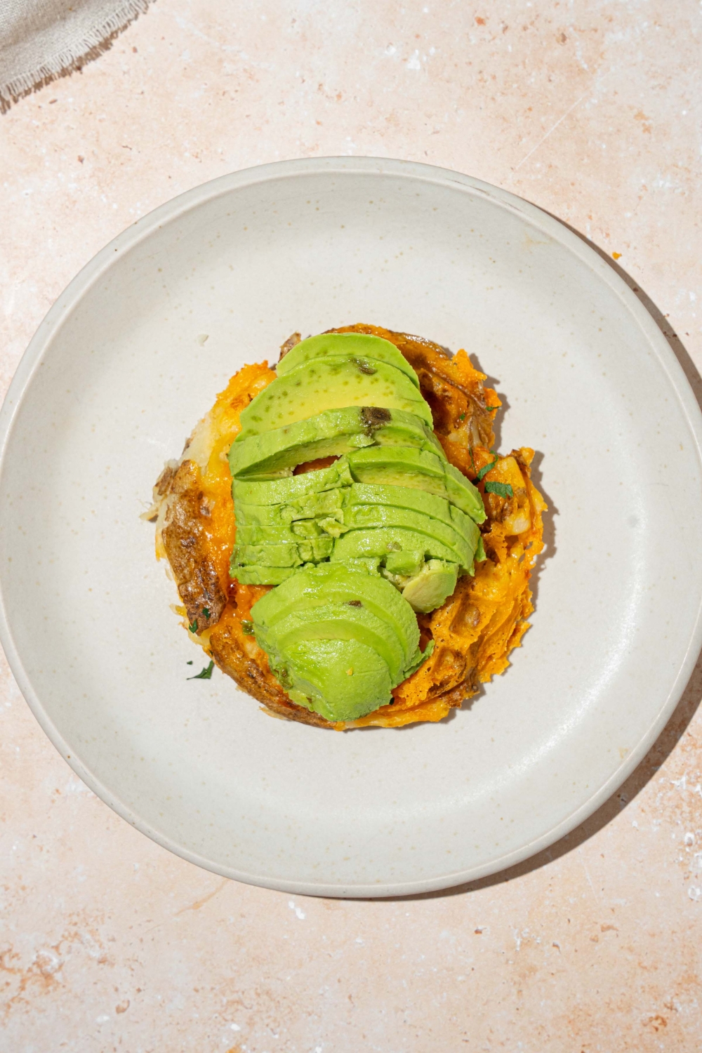 A white plate with a crispy potato waffle topped with sliced avocado. The plate is on a tan counter.