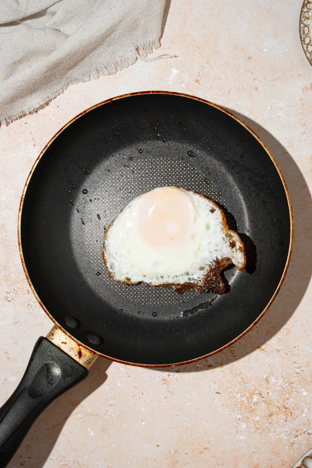 A skillet with a fried egg. The skillet is on a tan counter with a white cloth napkin.
