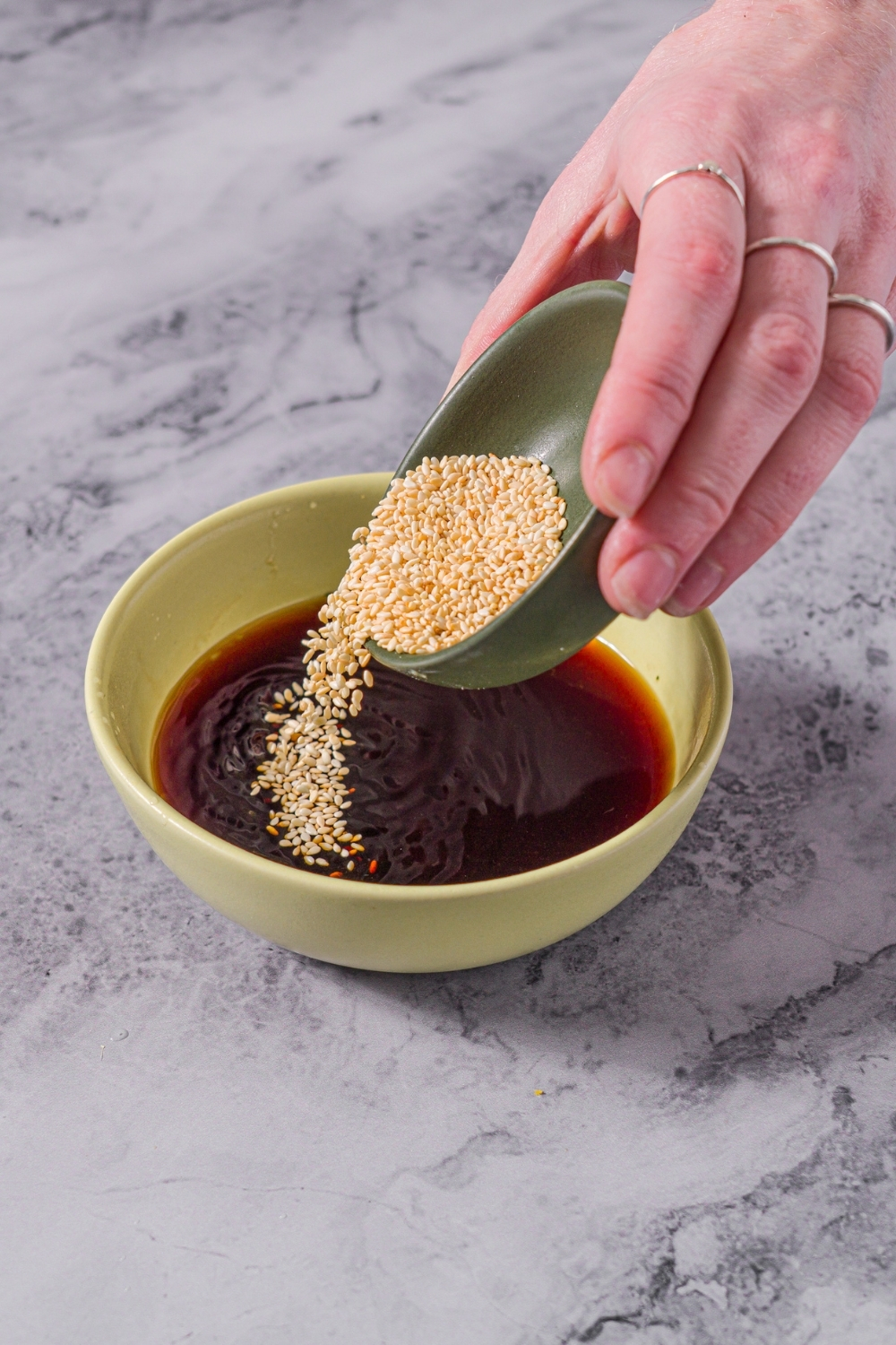 A small green bowl with soy sauce mixture for dumplings with a bowl of sesame seeds being added to the sauce. The bowl is on a marble counter.