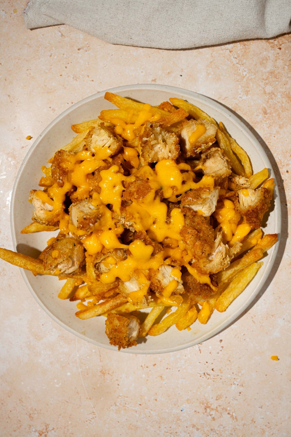 A white plate with fries topped with crispy chicken and melted cheese. The plate is on a tan counter with a white cloth napkin.