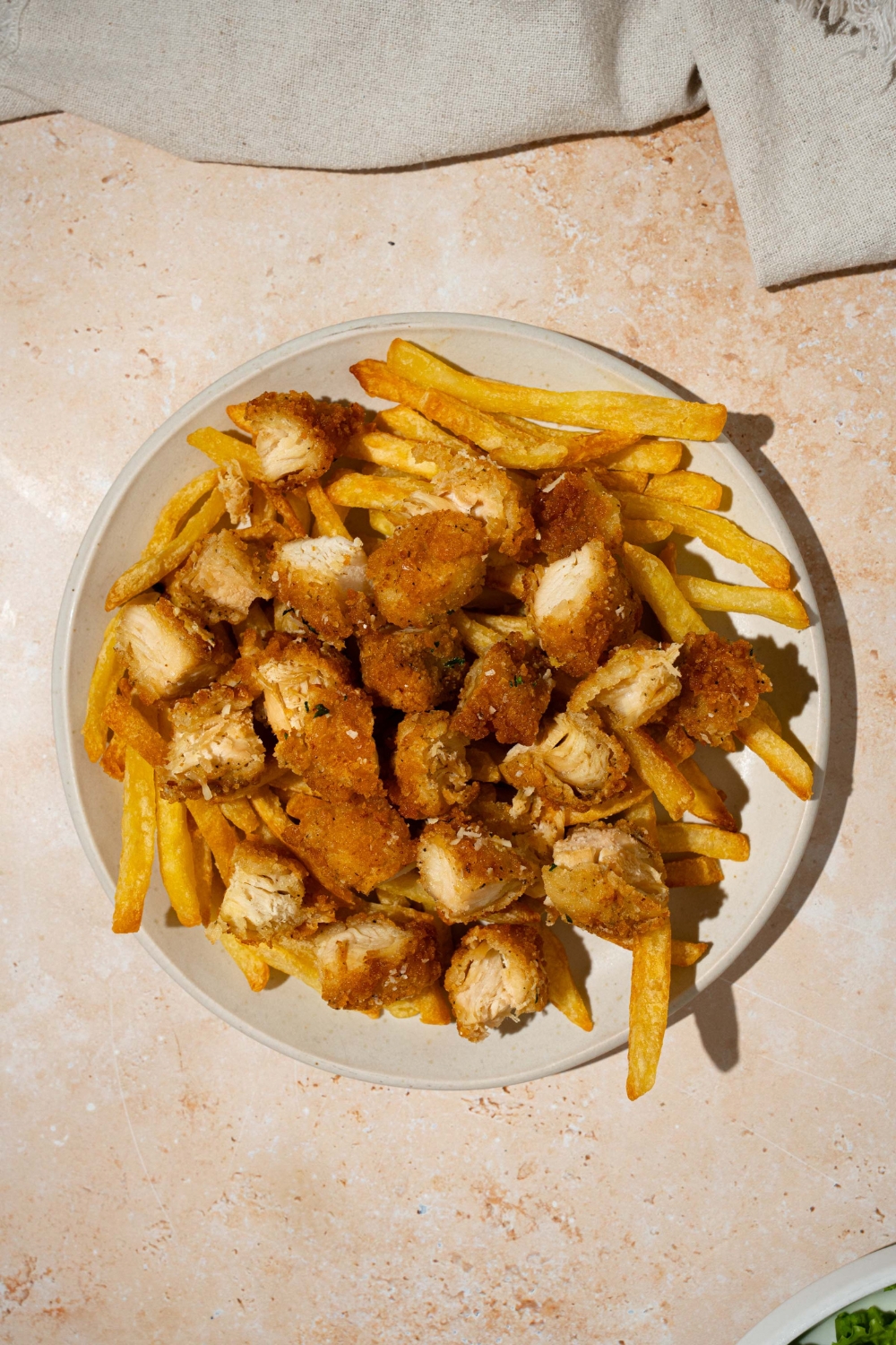 A white plate with fries topped with crispy chicken pieces. The plate is on a tan counter with a white cloth napkin.