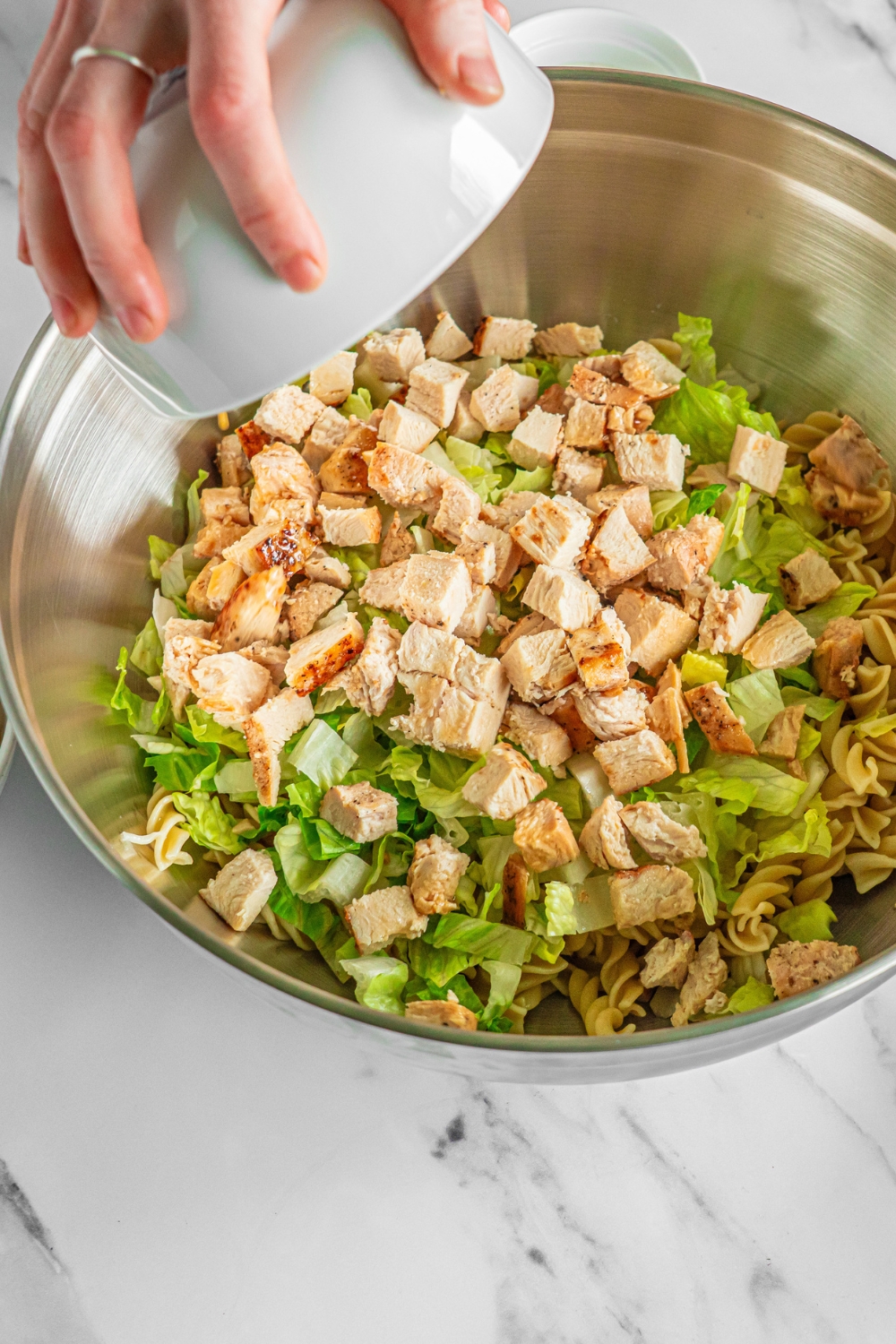A large bowl with rotini pasta, shredded lettuce, and a bowl of cubed chicken being added to the bowl. The bowl is on a marble counter.