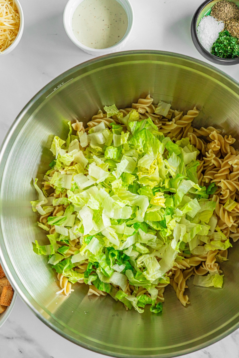 A large bowl with rotini pasta and shredded lettuce. The bowl is on a marble counter with small bowls of ingredients.