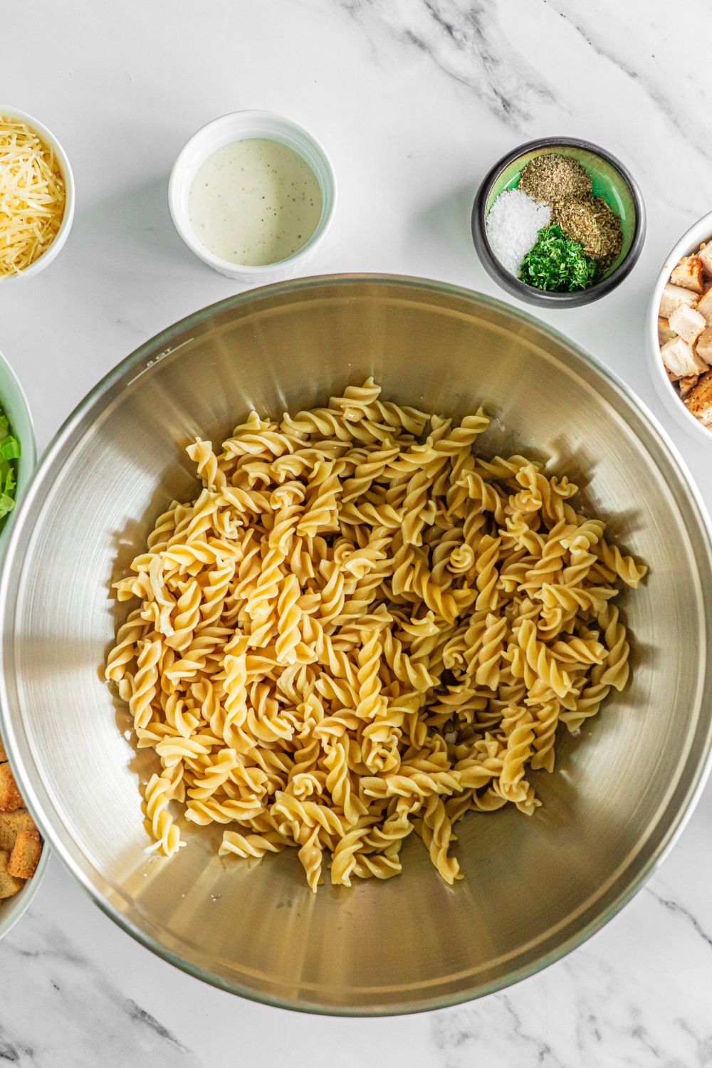 A large bowl with rotini pasta on a marble counter with small bowls of ingredients.