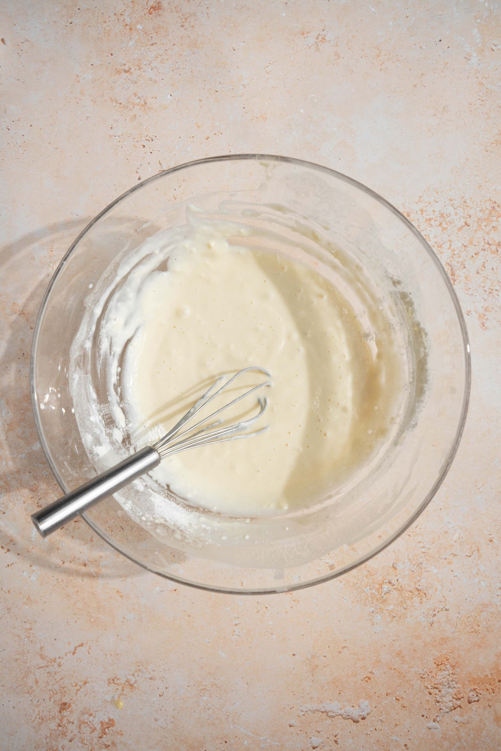 A glass bowl with a whisk mixing cinnamon roll pancake batter. The bowl is on a tan counter.