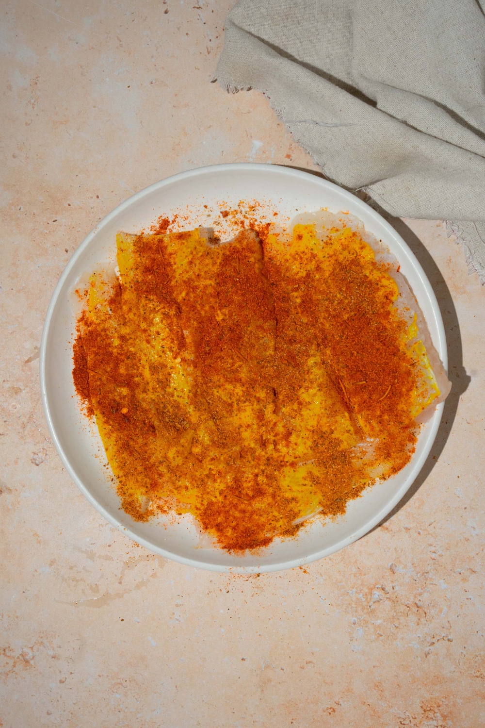 A white plate with fresh whiting coated in mustard and fish fry seasoning. The plate is on a tan counter.