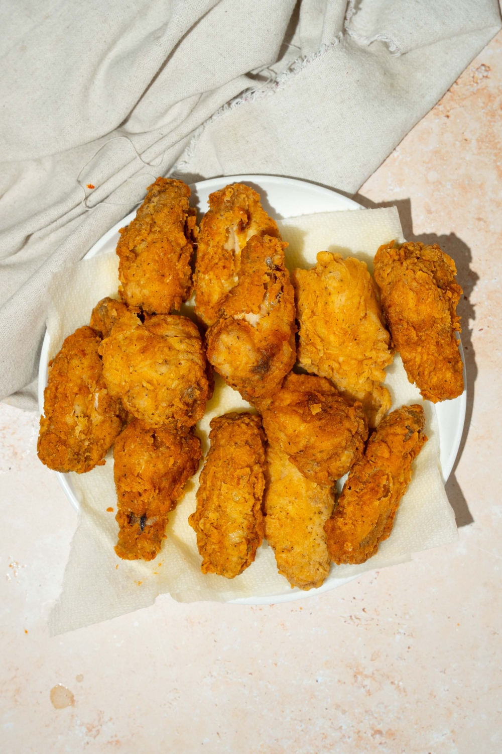 A white plate lined with paper towel with fried chicken wings. The plate is on a tan counter with a white cloth napkin.
