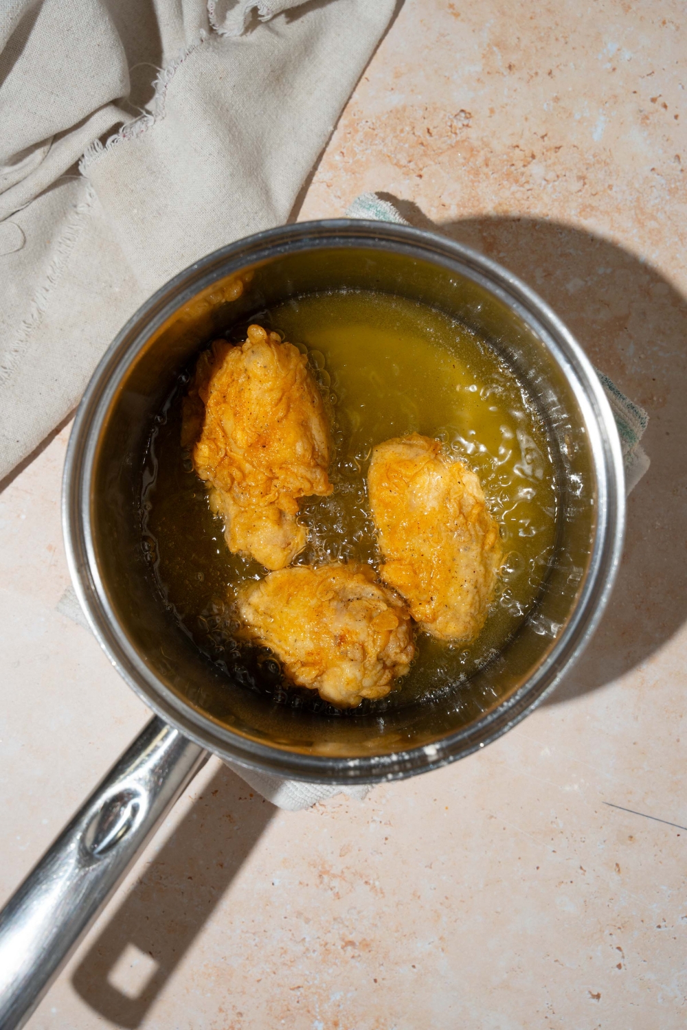A pot with chicken wings frying in oil. The pot is on a tan counter with a white cloth napkin.